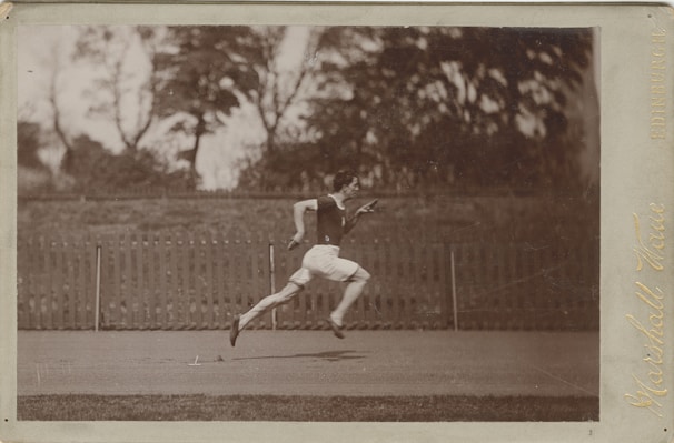 A monochrome photograph capturing a man in mid-sprint on an outdoor track. He is wearing a dark sleeveless top and light-colored shorts. The background shows a wooden fence and blurred, leafy trees.