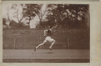 A monochrome photograph capturing a man in mid-sprint on an outdoor track. He is wearing a dark sleeveless top and light-colored shorts. The background shows a wooden fence and blurred, leafy trees.