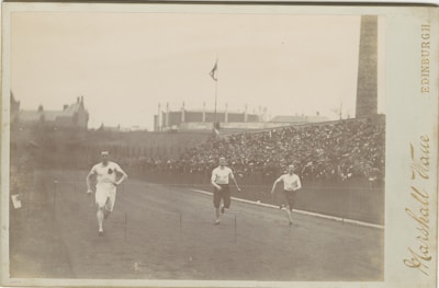Three athletes are sprinting on a track in an outdoor stadium, with one runner clearly leading. The stadium is filled with spectators, seated on bleachers that stretch along the side of the track. The background includes buildings and a tall chimney or tower. The photograph has a sepia tone, suggesting its historical nature.