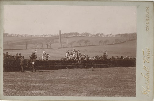 Several horse riders are jumping over a hedge in a vast open field, observed by a group of spectators. The background features rolling hills and sparse trees under a clear sky.