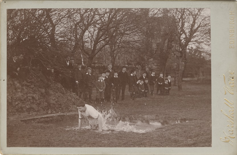 A group of boys dressed in formal clothing, likely from a bygone era, are gathered in a park setting with bare trees. One boy is kneeling in a small puddle of water, splashing around, while the others watch and seem amused. The scene appears to be playful and candid, capturing a moment of childhood mischief.