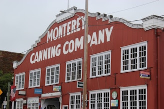 A historic red building with multiple white-paned windows and large white letters reading 'Monterey Canning Company.' The structure has a classic design with signs of various businesses along the street level, including a Starbucks and other local shops. An American flag is mounted on the left, with greenery visible in the background.