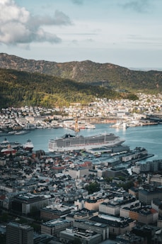 A bustling port city with a large cruise ship docked near the waterfront. The cityscape includes a mix of modern and classic architecture, surrounded by lush green hills under a partly cloudy sky.