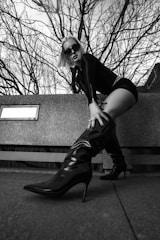 A person wearing a black outfit and sunglasses is posing with one leg extended forward, prominently displaying a high-heeled boot. The background features bare tree branches and a concrete structure, creating a stark and edgy atmosphere.