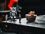 A figurine of a cat lifting a barbell is positioned on a wooden surface next to a toy mouse sitting in a wooden bucket. The setting has a playful and nostalgic atmosphere.