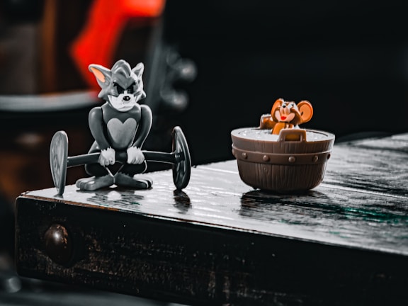 A figurine of a cat lifting a barbell is positioned on a wooden surface next to a toy mouse sitting in a wooden bucket. The setting has a playful and nostalgic atmosphere.