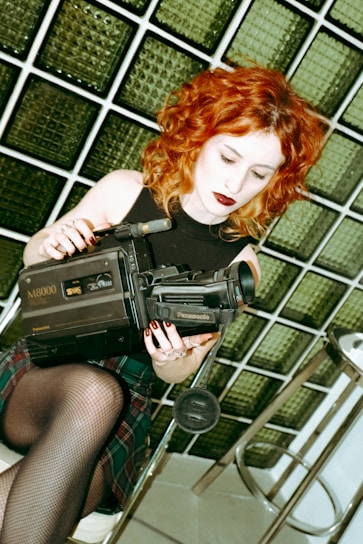 A person with curly red hair holding a vintage Panasonic M8000 video camera. They are sitting on a stool, wearing a black sleeveless top, plaid skirt, and fishnet stockings. The background consists of square glass tiles with a greenish tint.