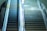 An escalator and a flight of stairs side by side. The escalator is on the left and appears to be stationary, with yellow caution signs at the edges. The stairs on the right are made of concrete or stone. The area is well-lit, with reflections visible on the surfaces.