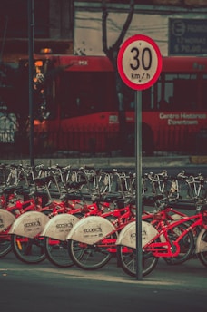 A row of red bicycles is lined up in a bike-sharing station, with a speed limit sign reading 30 km/h positioned prominently in the foreground. A red bus is visible in the background, partially obscured by trees and urban infrastructure.