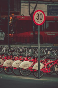 A row of red bicycles is lined up in a bike-sharing station, with a speed limit sign reading 30 km/h positioned prominently in the foreground. A red bus is visible in the background, partially obscured by trees and urban infrastructure.