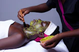 A person is lying down wrapped in a white towel while another person applies a green facial mask using a brush. The masked individual has a towel wrapped around their head and the setting suggests a spa or skincare treatment environment.
