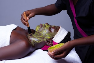 A person is lying down wrapped in a white towel while another person applies a green facial mask using a brush. The masked individual has a towel wrapped around their head and the setting suggests a spa or skincare treatment environment.