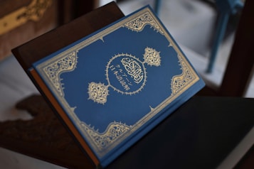 A beautifully designed, closed book with an intricate golden pattern and Arabic script on its blue cover, placed on a wooden stand.