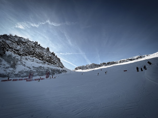 Snow-covered ski slope set against a mountainous backdrop, with a few skiers visible on the slope. Sparse trees line the edges of the mountains. The sky is a deep blue with streaks of clouds, and there are colorful banners along the sides of the slope.