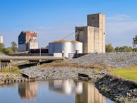 A large industrial facility with multiple silos and storage buildings sits alongside a body of water. The buildings have a weathered, rustic appearance with some areas of rust. A bridge spans over the water, leading to the facility. The surrounding area includes a rocky embankment and some grassy patches, with trees visible in the background under a clear blue sky.