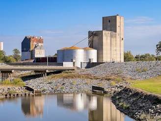A large industrial facility with multiple silos and storage buildings sits alongside a body of water. The buildings have a weathered, rustic appearance with some areas of rust. A bridge spans over the water, leading to the facility. The surrounding area includes a rocky embankment and some grassy patches, with trees visible in the background under a clear blue sky.