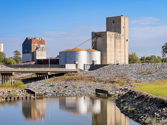 A large industrial facility with multiple silos and storage buildings sits alongside a body of water. The buildings have a weathered, rustic appearance with some areas of rust. A bridge spans over the water, leading to the facility. The surrounding area includes a rocky embankment and some grassy patches, with trees visible in the background under a clear blue sky.