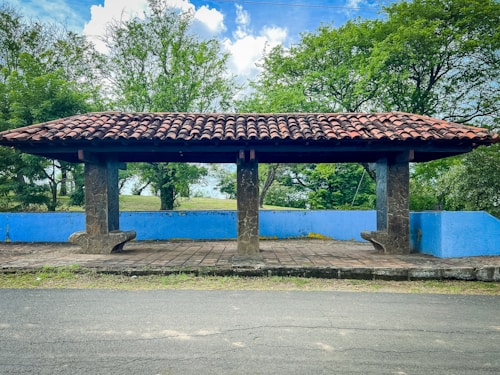 An outdoor shelter with a tiled roof stands on a paved area. It is supported by stone columns surrounded by greenery, including tall trees in the background. A blue painted low wall runs parallel to the shelter, adding a splash of color to the natural setting.