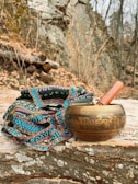 A brass singing bowl with intricate carvings sits on a stone surface, accompanied by a wooden mallet. Beside it, a colorful woven fabric with detailed patterns adds vibrancy to the scene. The background features a natural, rocky terrain with dry foliage and bare trees, suggesting a serene, outdoor setting.