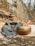 A brass singing bowl with intricate carvings sits on a stone surface, accompanied by a wooden mallet. Beside it, a colorful woven fabric with detailed patterns adds vibrancy to the scene. The background features a natural, rocky terrain with dry foliage and bare trees, suggesting a serene, outdoor setting.
