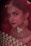 A woman adorned in traditional bridal jewelry and attire, featuring a red veil and intricate gold accessories. The face is partially covered by a delicate net fabric, and she wears a large nose ring and elaborate earrings. Her makeup is elegantly done, highlighting her eyes and lips.