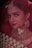 A woman adorned in traditional bridal jewelry and attire, featuring a red veil and intricate gold accessories. The face is partially covered by a delicate net fabric, and she wears a large nose ring and elaborate earrings. Her makeup is elegantly done, highlighting her eyes and lips.