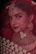 A woman adorned in traditional bridal jewelry and attire, featuring a red veil and intricate gold accessories. The face is partially covered by a delicate net fabric, and she wears a large nose ring and elaborate earrings. Her makeup is elegantly done, highlighting her eyes and lips.