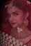 A woman adorned in traditional bridal jewelry and attire, featuring a red veil and intricate gold accessories. The face is partially covered by a delicate net fabric, and she wears a large nose ring and elaborate earrings. Her makeup is elegantly done, highlighting her eyes and lips.