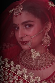 A woman adorned in traditional bridal jewelry and attire, featuring a red veil and intricate gold accessories. The face is partially covered by a delicate net fabric, and she wears a large nose ring and elaborate earrings. Her makeup is elegantly done, highlighting her eyes and lips.