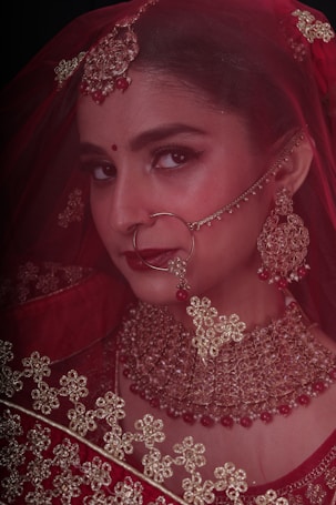 A woman adorned in traditional bridal jewelry and attire, featuring a red veil and intricate gold accessories. The face is partially covered by a delicate net fabric, and she wears a large nose ring and elaborate earrings. Her makeup is elegantly done, highlighting her eyes and lips.
