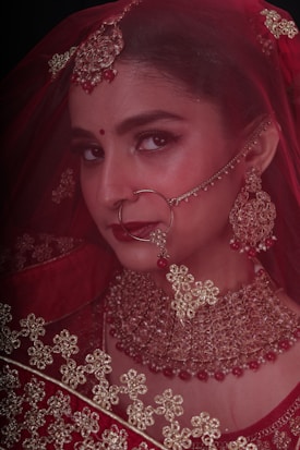 A woman adorned in traditional bridal jewelry and attire, featuring a red veil and intricate gold accessories. The face is partially covered by a delicate net fabric, and she wears a large nose ring and elaborate earrings. Her makeup is elegantly done, highlighting her eyes and lips.