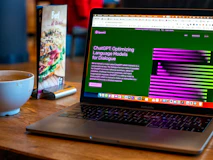 A laptop displaying a website about language models is set on a wooden table. A coffee cup is nearby, next to a menu stand featuring a beef dish advertisement.
