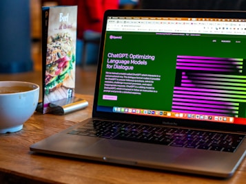 A laptop displaying a website about language models is set on a wooden table. A coffee cup is nearby, next to a menu stand featuring a beef dish advertisement.