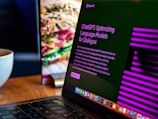 A laptop displays a screen with the title 'ChatGPT: Optimizing Language Models for Dialogue', accompanied by descriptive text. The background shows a blurred image of a sandwich, and there's a white cup on the wooden table next to the laptop.
