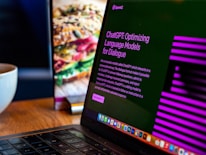 A laptop displays a screen with the title 'ChatGPT: Optimizing Language Models for Dialogue', accompanied by descriptive text. The background shows a blurred image of a sandwich, and there's a white cup on the wooden table next to the laptop.