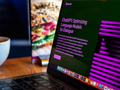 A laptop displays a screen with the title 'ChatGPT: Optimizing Language Models for Dialogue', accompanied by descriptive text. The background shows a blurred image of a sandwich, and there's a white cup on the wooden table next to the laptop.