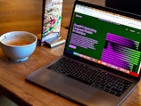 A laptop displaying a webpage about optimizing language models rests on a wooden table. To the left of the laptop is a white cup containing coffee, with remnants of foam around the edges. A colorful laminated menu stand with a sandwich picture is positioned behind the cup.