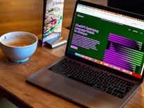 A laptop displaying a webpage about optimizing language models rests on a wooden table. To the left of the laptop is a white cup containing coffee, with remnants of foam around the edges. A colorful laminated menu stand with a sandwich picture is positioned behind the cup.