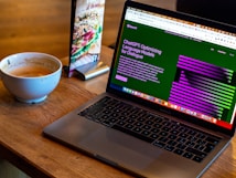 A laptop displaying a webpage about optimizing language models rests on a wooden table. To the left of the laptop is a white cup containing coffee, with remnants of foam around the edges. A colorful laminated menu stand with a sandwich picture is positioned behind the cup.