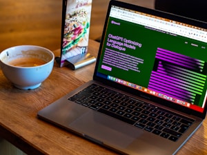 A laptop displaying a webpage about optimizing language models rests on a wooden table. To the left of the laptop is a white cup containing coffee, with remnants of foam around the edges. A colorful laminated menu stand with a sandwich picture is positioned behind the cup.