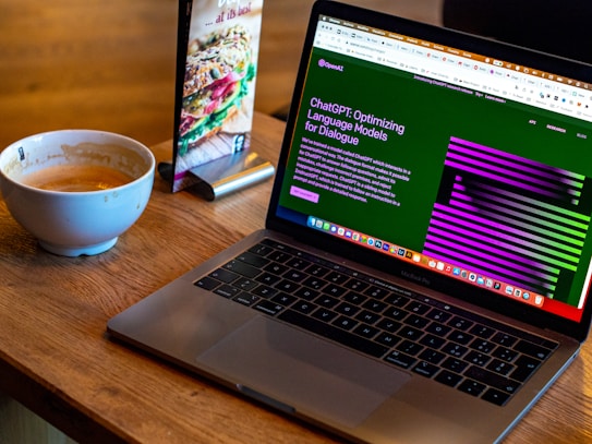 A laptop displaying a webpage about optimizing language models rests on a wooden table. To the left of the laptop is a white cup containing coffee, with remnants of foam around the edges. A colorful laminated menu stand with a sandwich picture is positioned behind the cup.