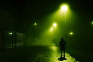A lone figure in a dark hoodie walking under a streetlamp casting long shadows.