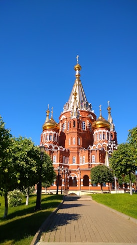 A stunning orthodox church with red brick walls and golden domes stands elegantly under a clear blue sky. The architecture features intricate designs, with several spires topped by gilded crosses. Surrounding the building are lush green trees and a paved walkway leading up to it.