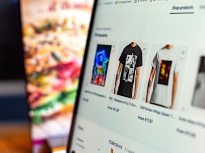The focus is on a computer screen displaying an online store featuring T-shirts with various graphic designs. The left side of the image is blurred, showing indistinct colors and shapes, possibly a menu or food items.