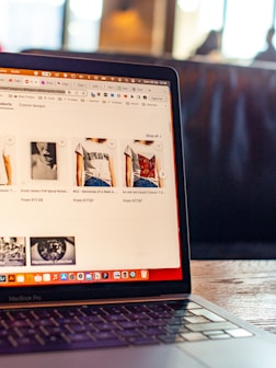 The image features an open laptop displaying an online shopping website. The screen shows various t-shirts with artistic designs, priced in euros. The setting appears to be in a cafe or a relaxed workspace, with blurred figures and warm lighting in the background.