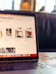The image features an open laptop displaying an online shopping website. The screen shows various t-shirts with artistic designs, priced in euros. The setting appears to be in a cafe or a relaxed workspace, with blurred figures and warm lighting in the background.