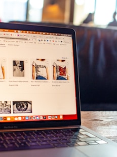 The image features an open laptop displaying an online shopping website. The screen shows various t-shirts with artistic designs, priced in euros. The setting appears to be in a cafe or a relaxed workspace, with blurred figures and warm lighting in the background.