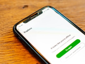 A smartphone with a cracked screen displays a message indicating that it is offline with options to try again or read help articles. The device is resting on a wooden surface.