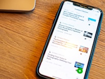 A smartphone displaying a list of articles with their titles, authors, and reading times is placed on a wooden surface. The phone screen is illuminated, and a part of a laptop is visible in the background.