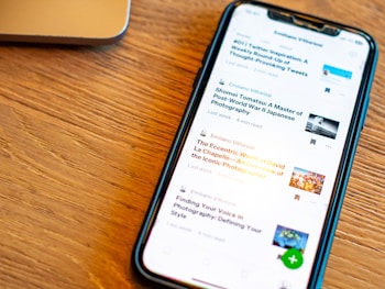 A smartphone displaying a list of articles with their titles, authors, and reading times is placed on a wooden surface. The phone screen is illuminated, and a part of a laptop is visible in the background.